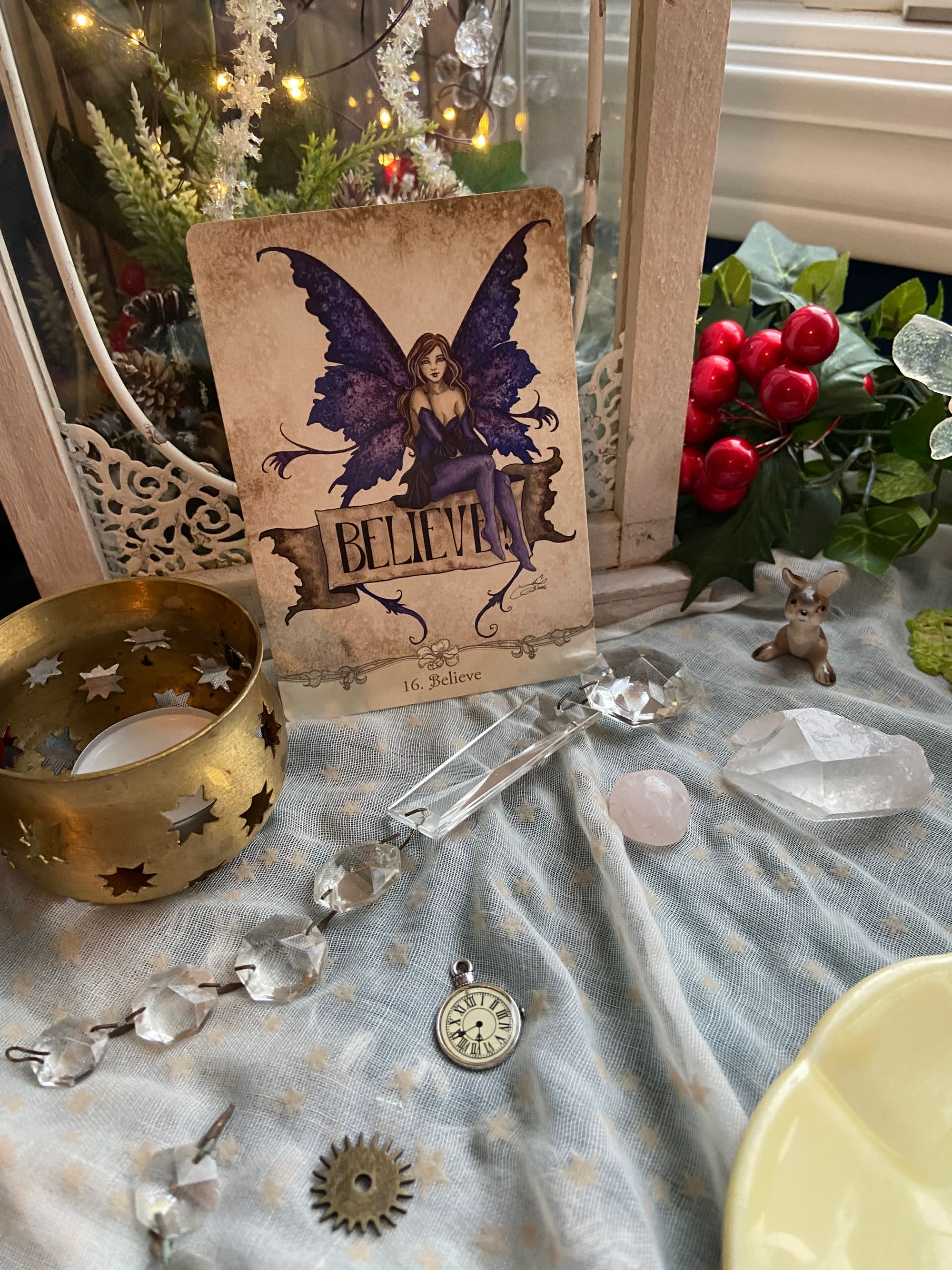 a table covered in greenery, red berries, clear crystals, a gold candle, a lantern, and a fairy oracle card